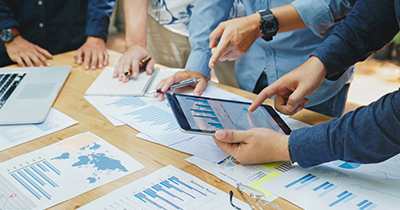 A group of people leaning over a desk with statistics displayed on tablet and documents.